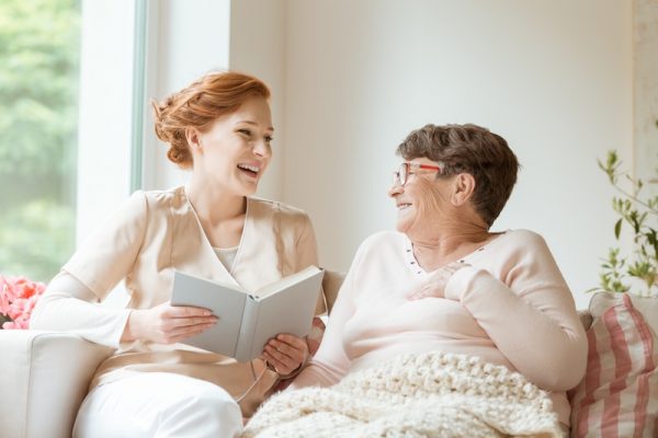 Happy nurse reading a funny book to her elderly patient in a private nursing home